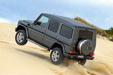 Mercedes G-Wagen climbs sand dune, spraying sand.