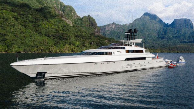 A superyacht floats near forested shore, mountains.