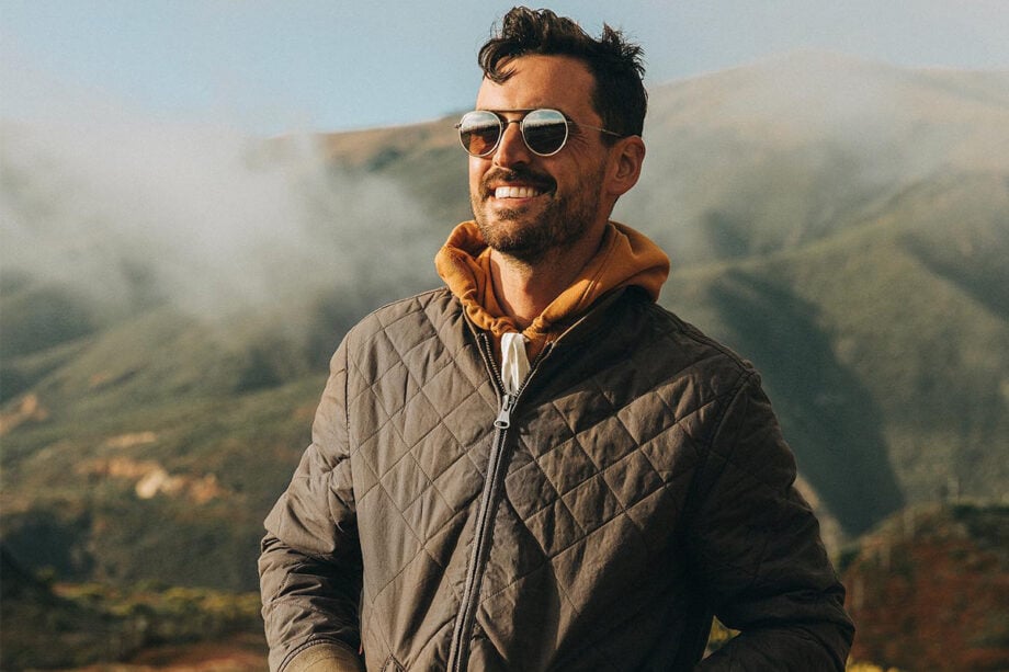 Man in designer jacket, smiling amidst foggy hills.